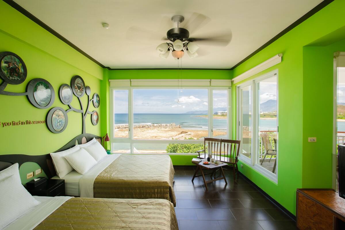 Una habitación con dos camas, ventilador de techo y ventanas amplias con vista a la playa.