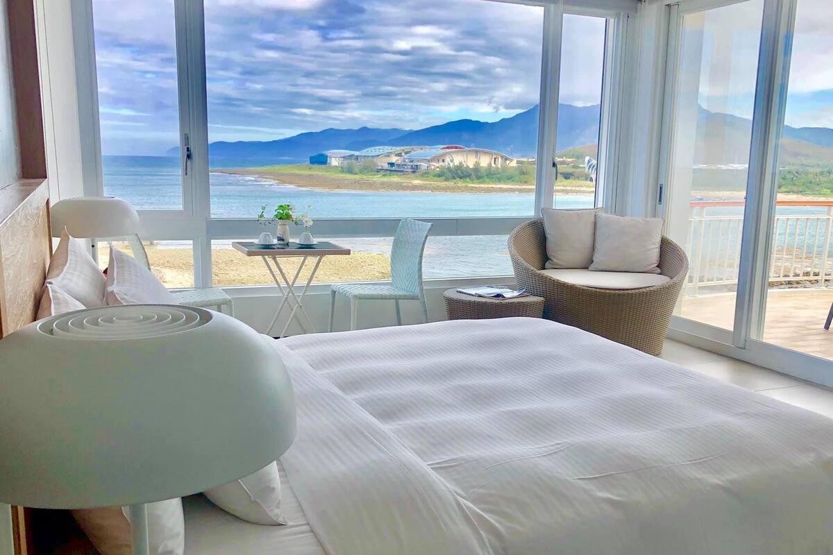 Un dormitorio con una cama grande, una silla, una mesita y vistas al mar y las montañas.