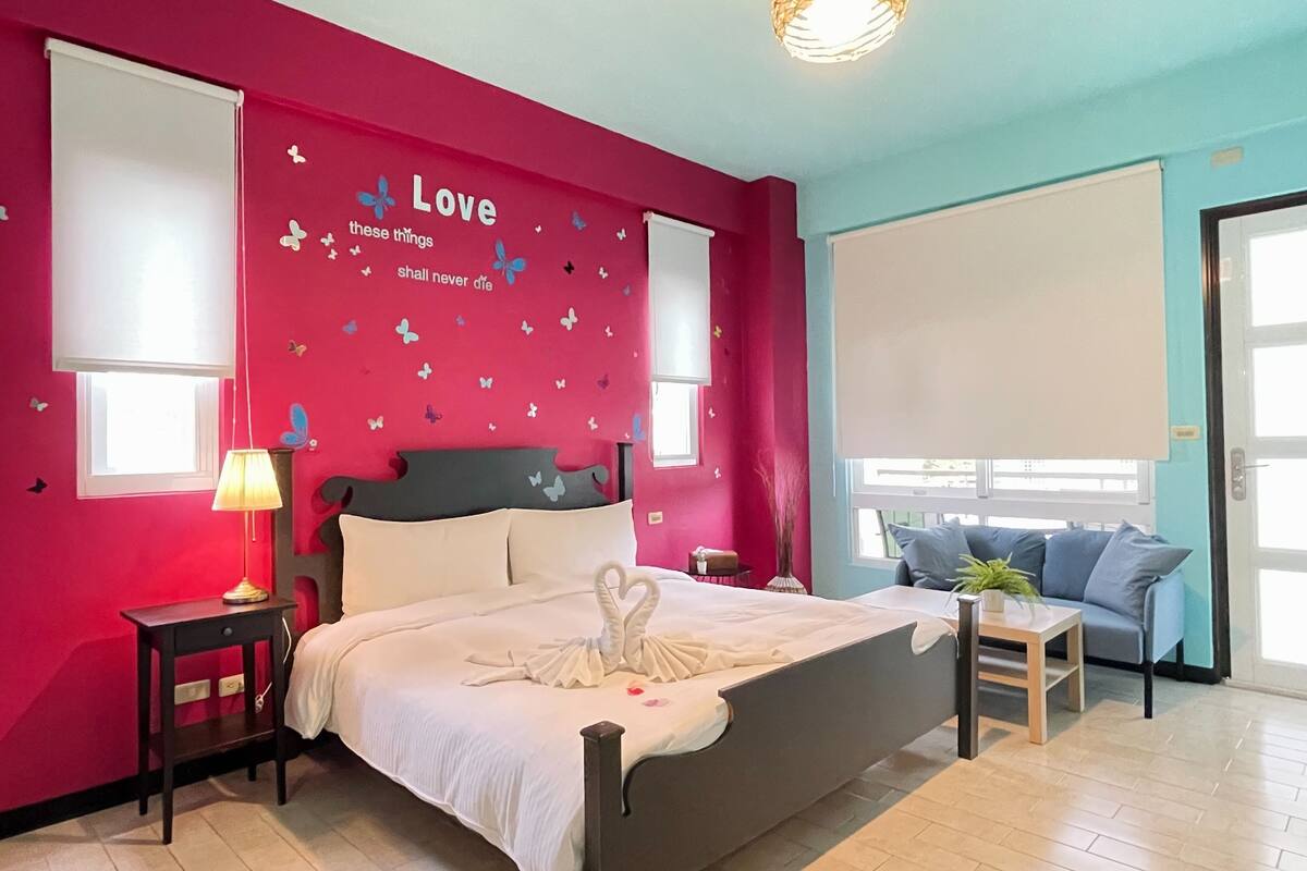Un dormitorio con cama, mesita de noche con lámpara, pared decorada con adhesivos de mariposas y ventana con vistas.