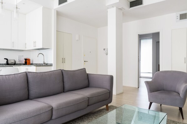 A modern living room with a grey sofa, a glass coffee table, and a grey armchair. There is a kitchen area with white cabinets and a black kettle on the counter.