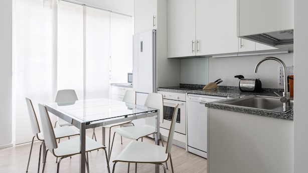Une cuisine moderne avec des meubles blancs, un plan de travail en granit, un évier en acier inoxydable et une table à manger en verre, assortie de chaises blanches.