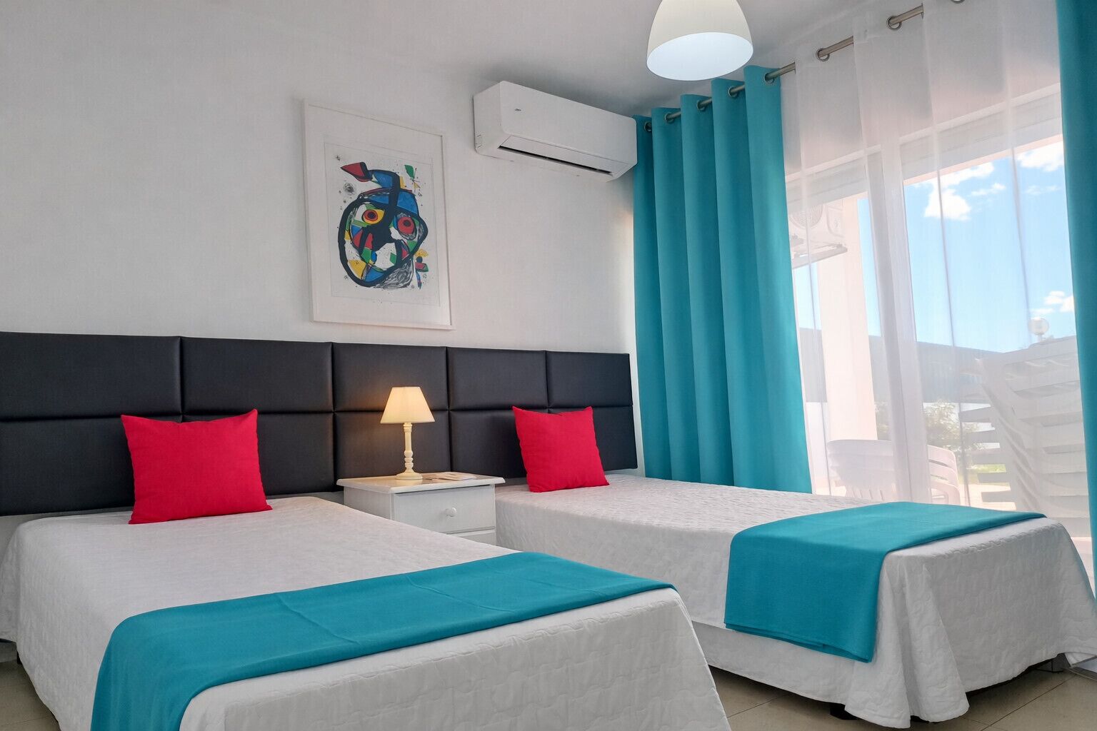 A hotel room with two beds, a nightstand with a lamp, a ceiling fan, and a large window with curtains.