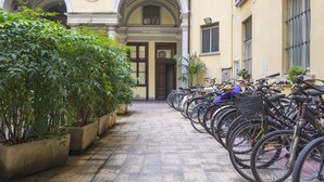 A row of bicycles parked in front of a building with a tiled floor and potted plants on the left.