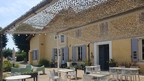 Un patio al aire libre con mesas y sillas de celosía blanca, un edificio amarillo con contraventanas y plantas en macetas.