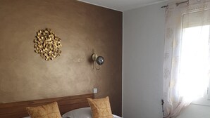 Una cama bien tendida con una manta color beige y dos almohadas estampadas, un aplique de pared y una pieza decorativa de arte con pan de oro.