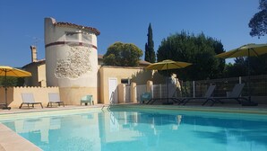 Una piscina con sillas de descanso y sombrillas, una torre de piedra y un área cercada con árboles.