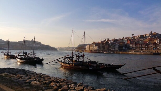 Un puerto con barcos amarrados, un rompeolas de piedra y un paisaje urbano con edificios al fondo.