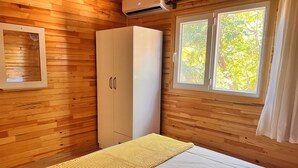 A wooden cabin room with a bed, a wardrobe, and a window with a view of trees.