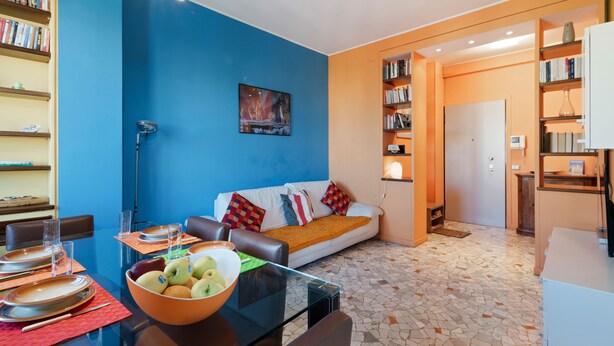 Uma sala de estar moderna com uma parede azul, uma mesa de centro de vidro, um sofá com almofadas coloridas e uma estante cheia de livros.