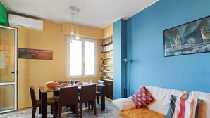 Uma sala de jantar com mesa e cadeiras, um sofá com almofadas, uma estante de livros e um quadro na parede.