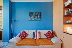A room with a blue wall, a white sofa with patterned cushions, a framed picture of wolves and the aurora borealis, and a bookshelf with books. - Flatty Apartments - Transiti (Milan)