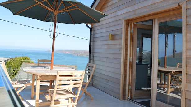 Eine Holzterrasse mit Tisch und Stühlen, einem grünen Sonnenschirm und einer Glastür, die auf einen Balkon mit Meerblick führt.