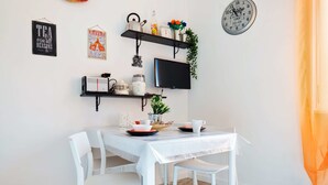 A dining area with a white table, white chairs, a flat-screen TV, and wall-mounted shelves with kitchen items and decorations. - Flatty Apartments - Alserio (Milan)