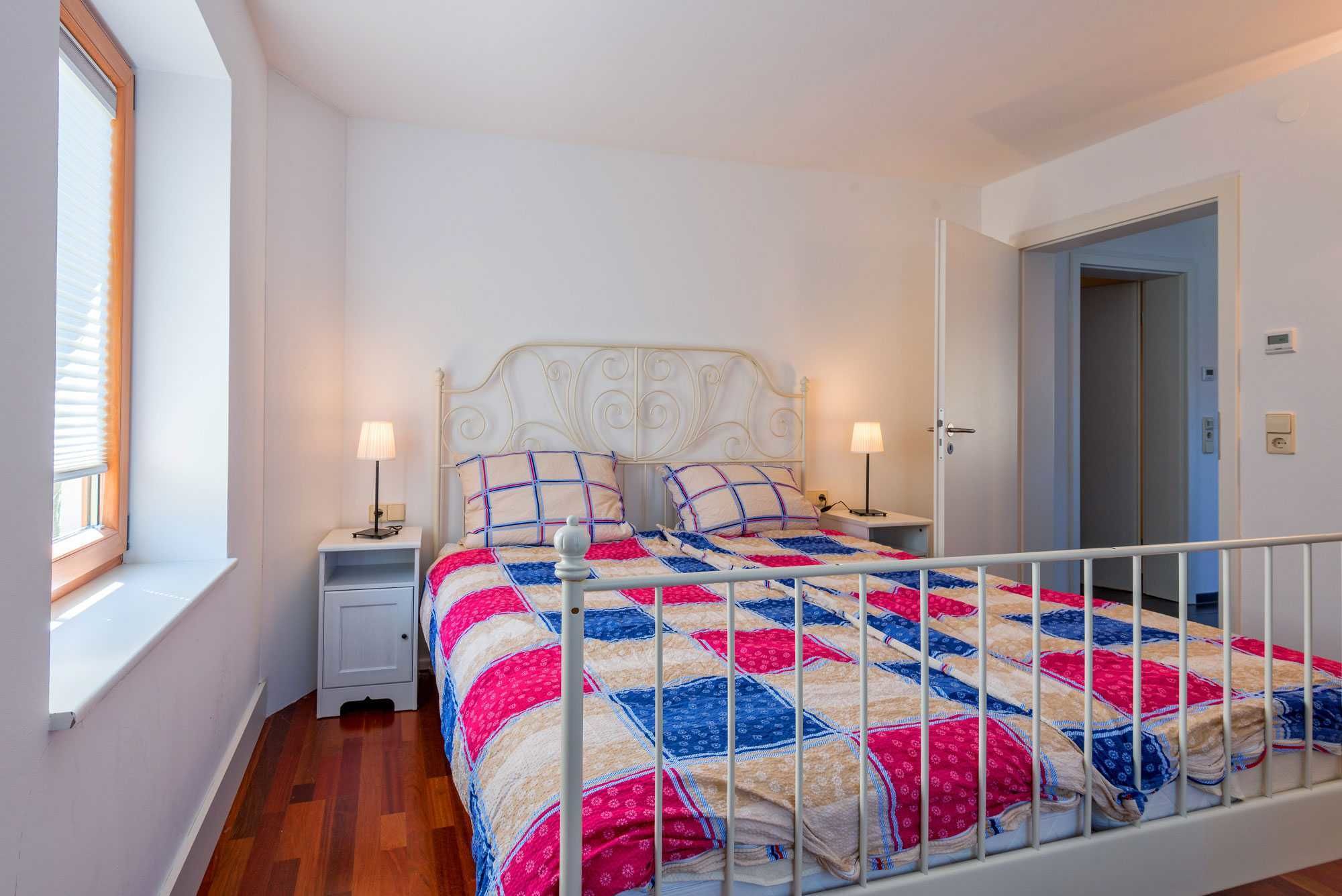 A bedroom with a bed, two bedside tables, a window with blinds, a door, and a white railing.