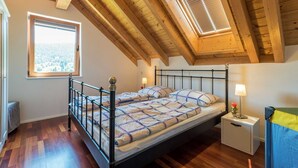 A bedroom with a sloped ceiling, a bed with a metal frame, two bedside tables with lamps, and a window with a view of a snowy landscape. - Wellness Ferienhaus Fronwald (Alpirsbach)
