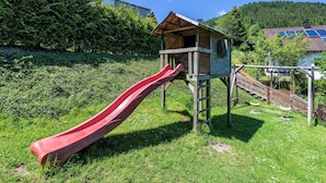 A wooden playhouse with a red slide, surrounded by greenery and a house with solar panels in the background. - Wellness Ferienhaus Fronwald (Alpirsbach)