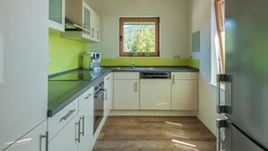 A modern kitchen with white cabinets, a green countertop, a built-in dishwasher, and a window with a view of trees. - Wellness Ferienhaus Fronwald (Alpirsbach)