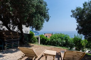 A coastal view with wooden lounge chairs and a table, overlooking the sea and a distant building. - Guesthouse Oliva (Dubrovnik)