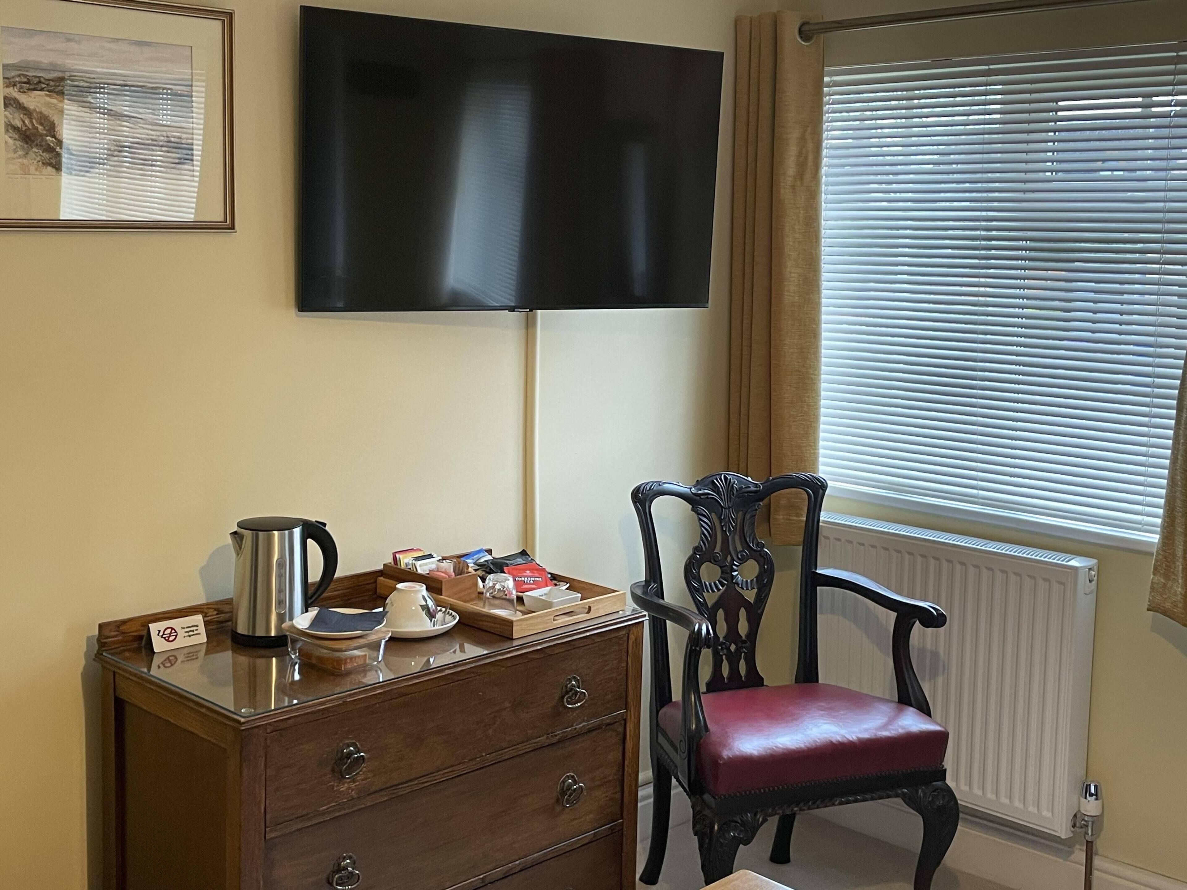 A room with a wooden dresser, a red leather chair, a flat-screen TV mounted on the wall, and a window with blinds.
