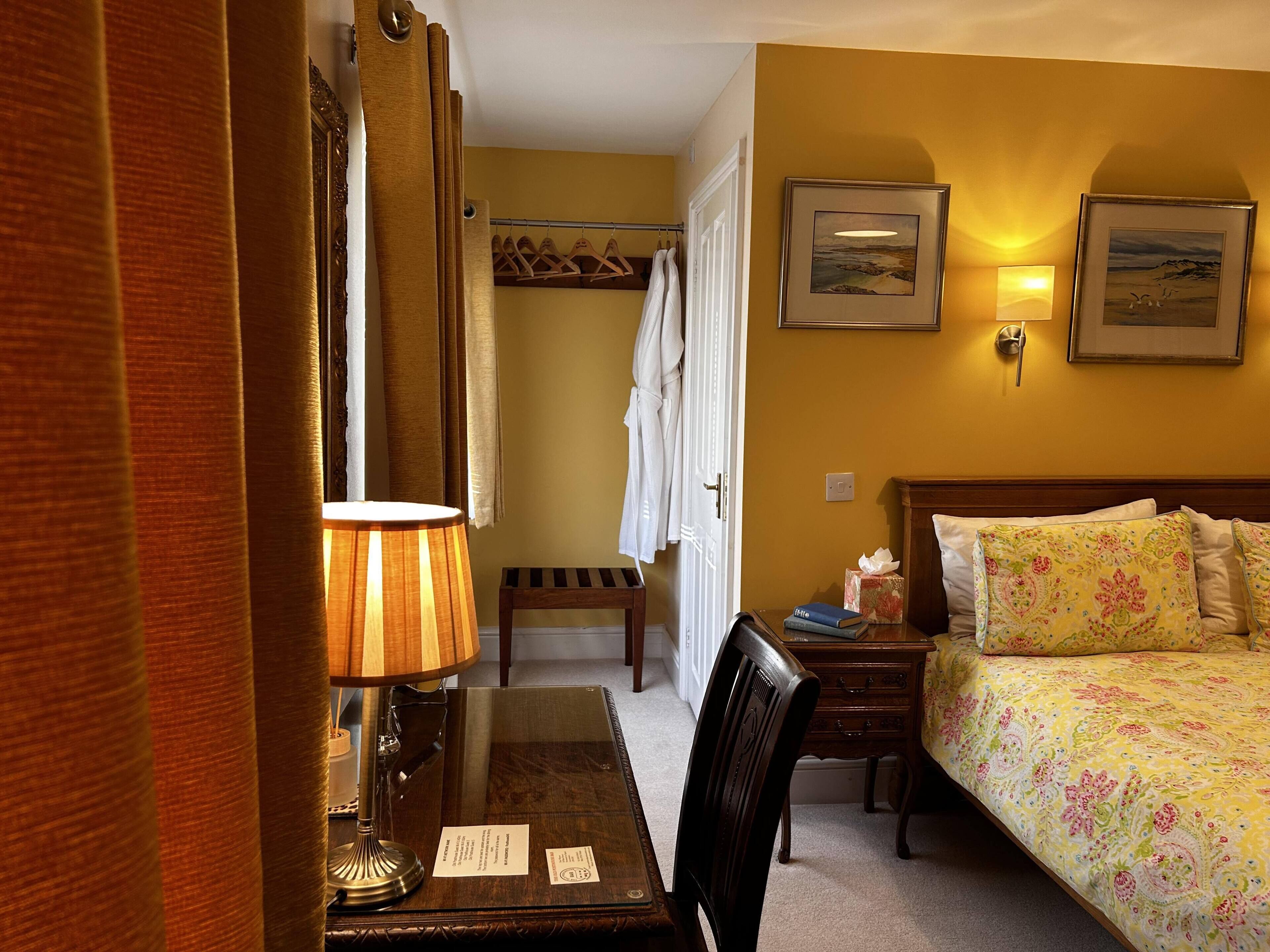 A hotel room with a bed, a desk with a lamp, a chair, a bedside table, a coat rack, and two framed pictures on the wall.