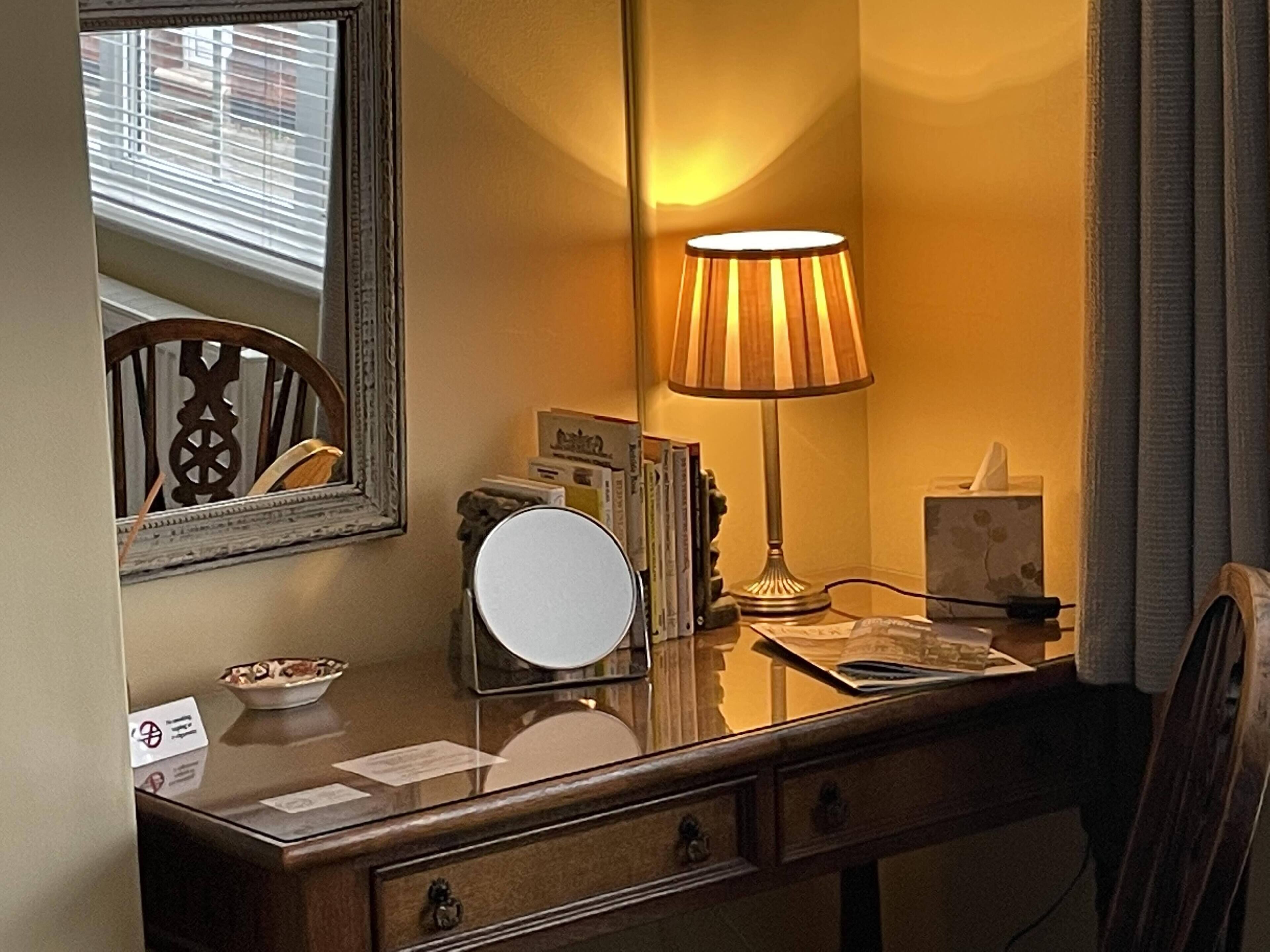 Habitación de hotel con un escritorio de madera, un espejo, una lámpara, un frutero y una ventana con persianas.