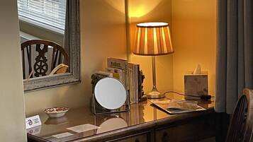 A hotel room with a wooden desk, a mirror, a lamp, a bowl of fruit, and a window with blinds.