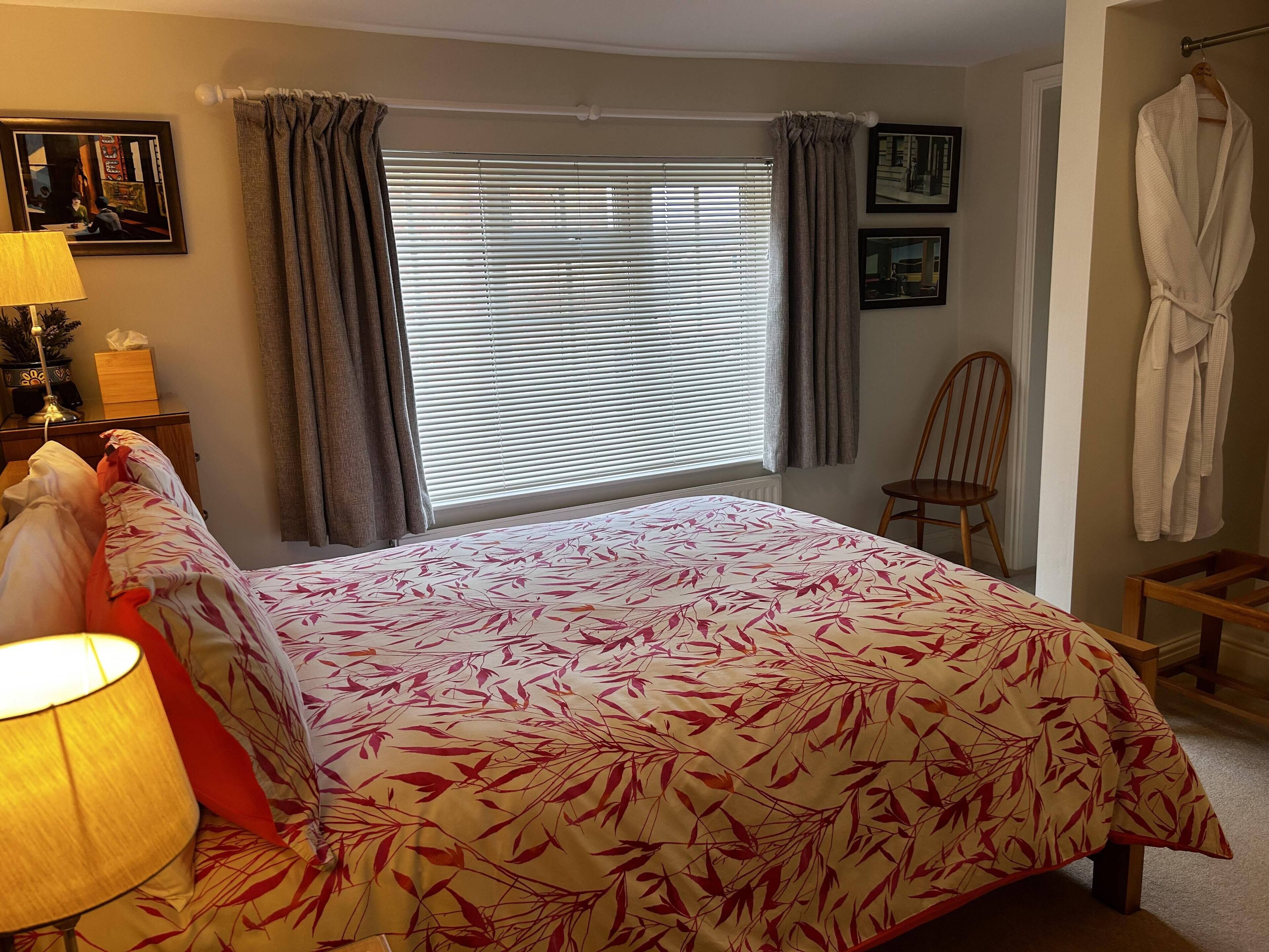 Una cama bien hecha con una colcha floral, una lámpara de noche, una silla y una bata colgada en la pared.