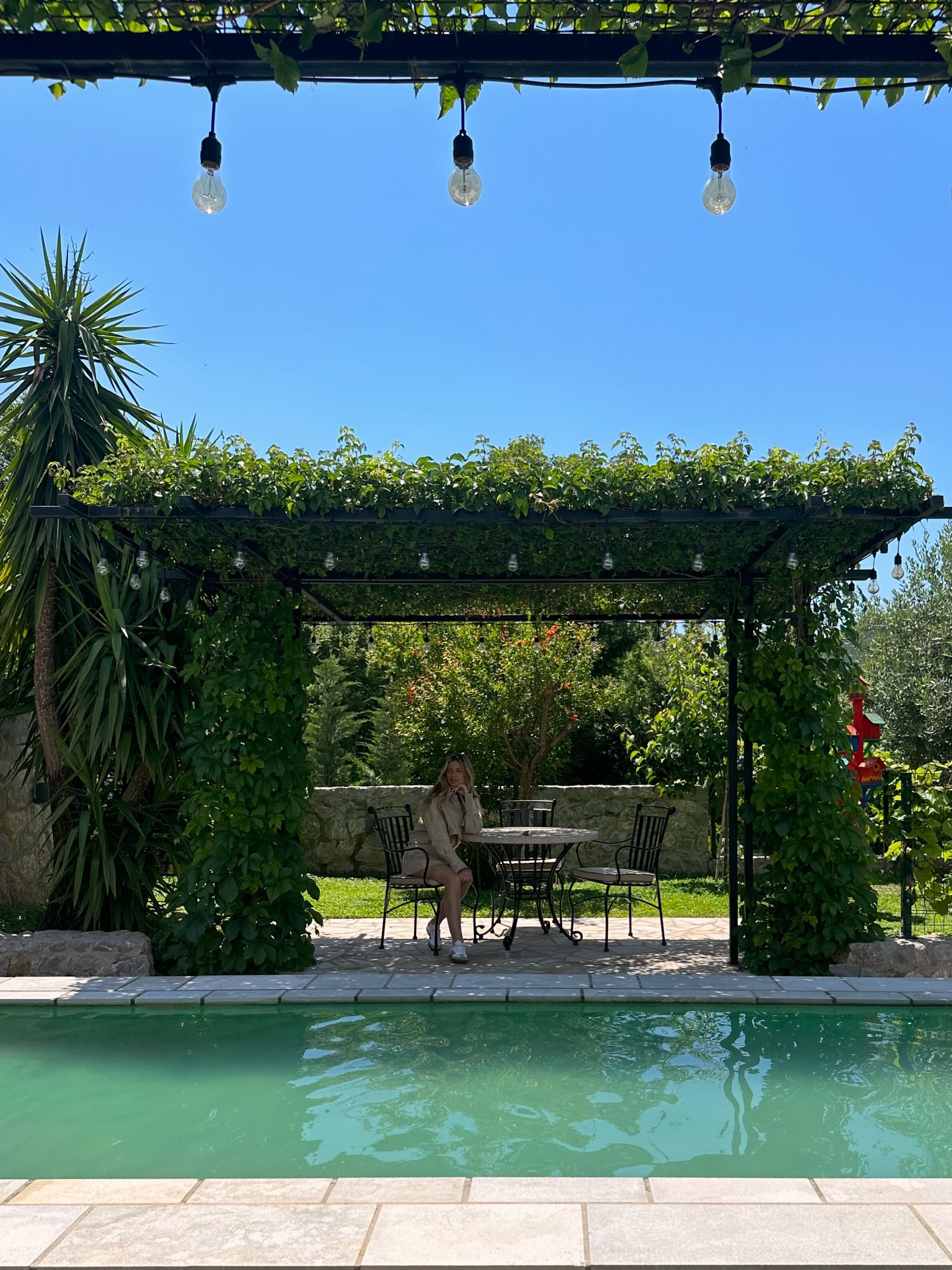 Uma área de estar coberta ao ar livre com piscina, rodeada por vegetação e um céu azul claro.