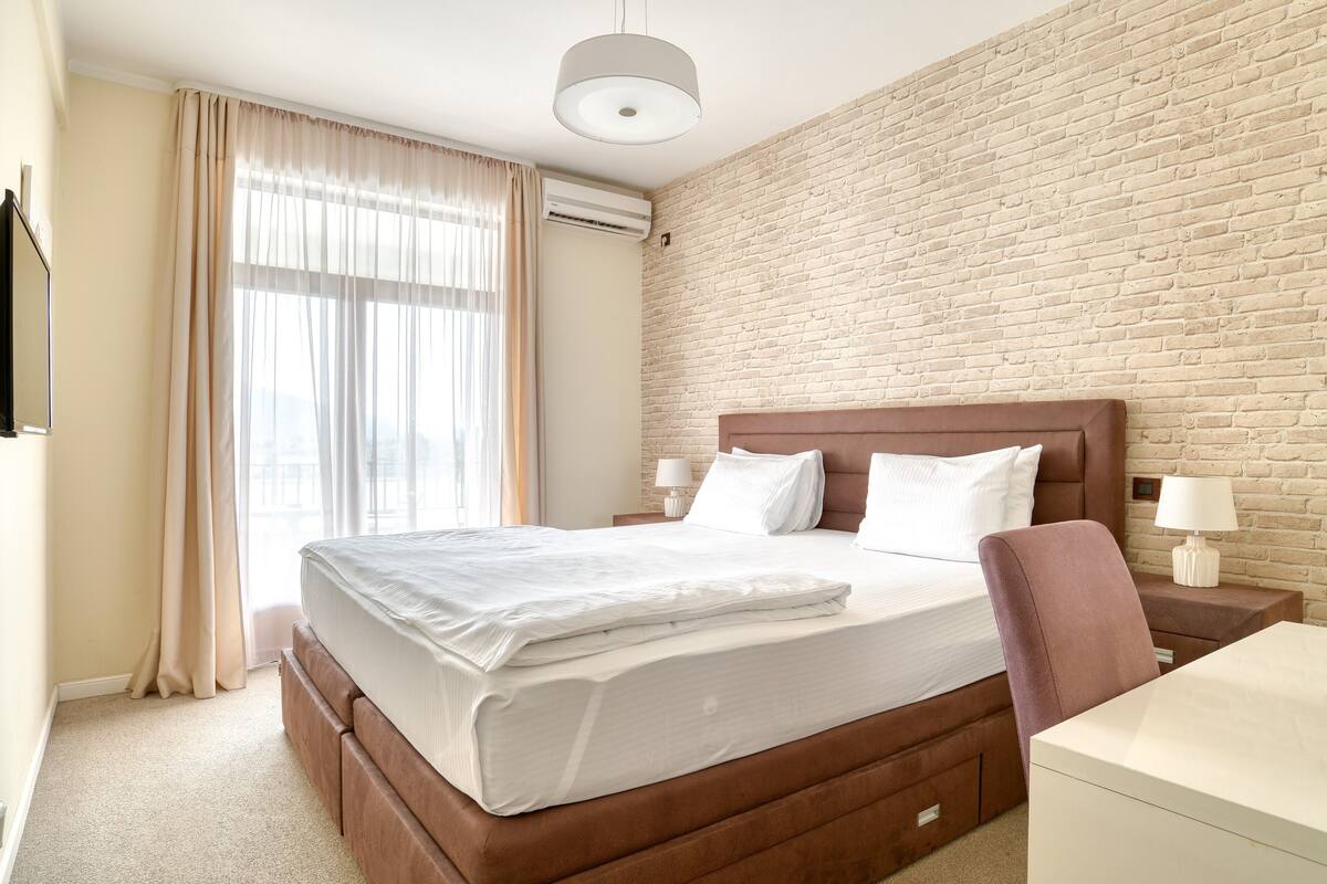 A modern hotel room with a brick wall, a large bed with white linens, a wooden headboard, two bedside tables with lamps, and a view of the city through large windows.