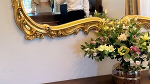 Una habitación con un espejo grande y ornamentado, una planta en maceta con flores y una ventana con cortinas.
