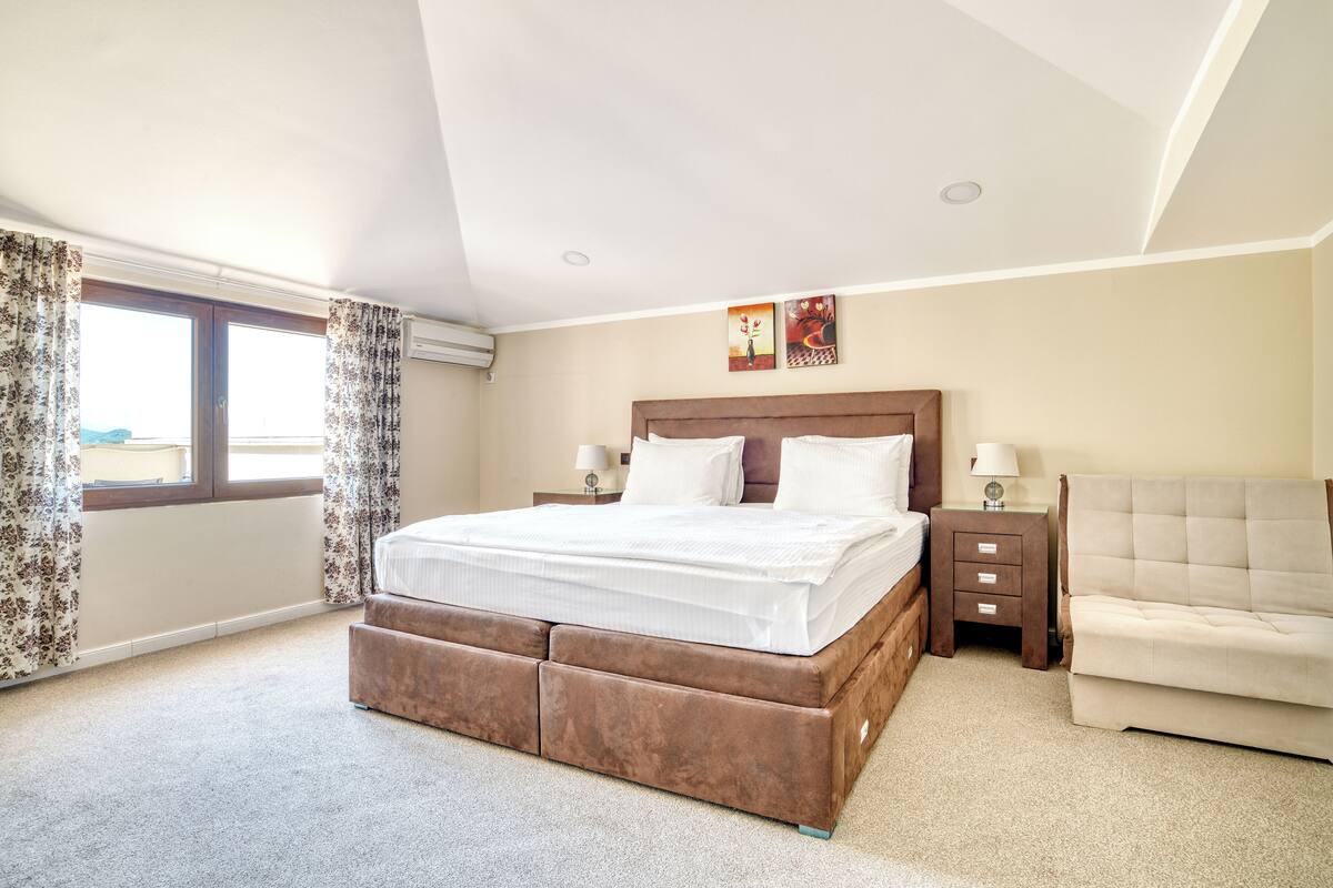 A bedroom with a large bed, a nightstand, a chair, and a window with curtains.