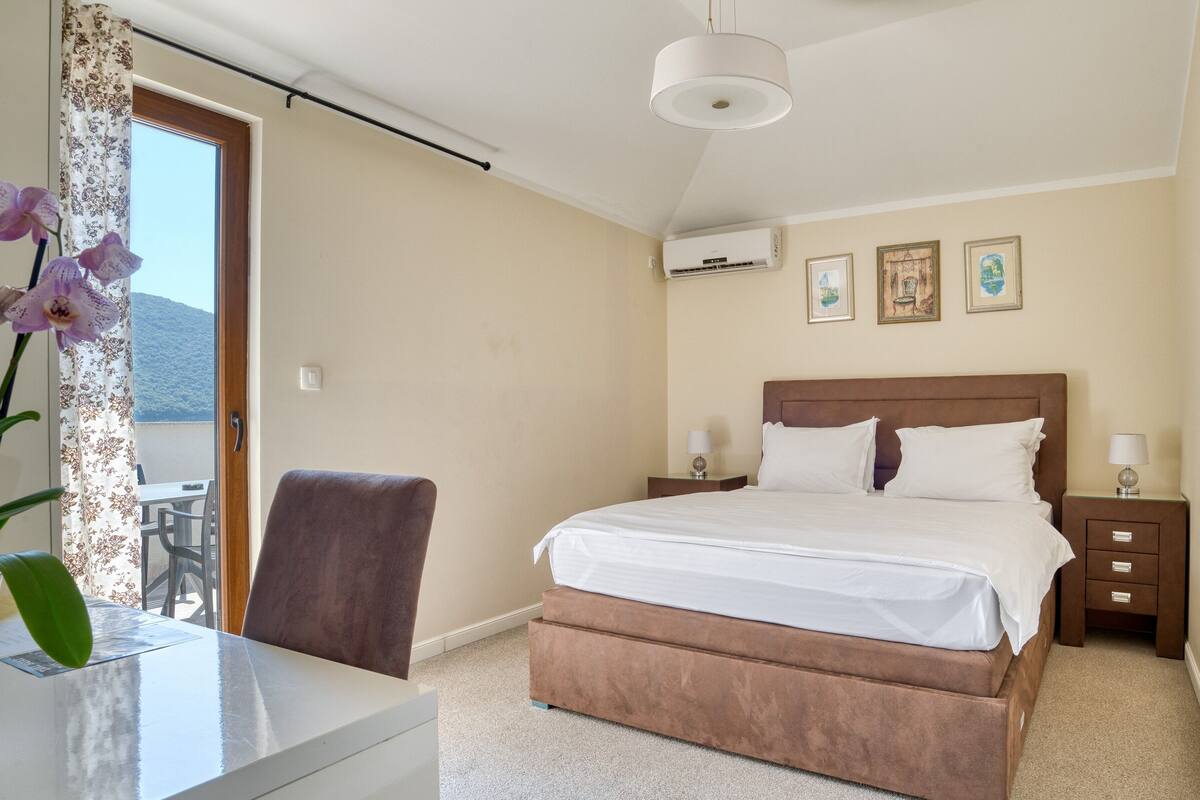 A bedroom with a bed, bedside tables, a chair, a desk, and a view of the outdoors.