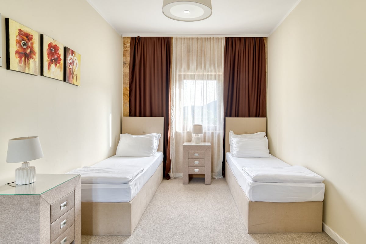 A hotel room with two single beds, a bedside table with a lamp, a small dresser, and a window with curtains.