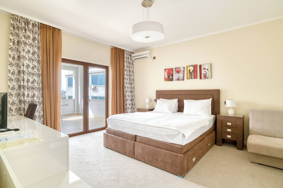 A hotel room with a bed, bedside tables, a desk, and a chair. There is a sliding glass door leading to a balcony.