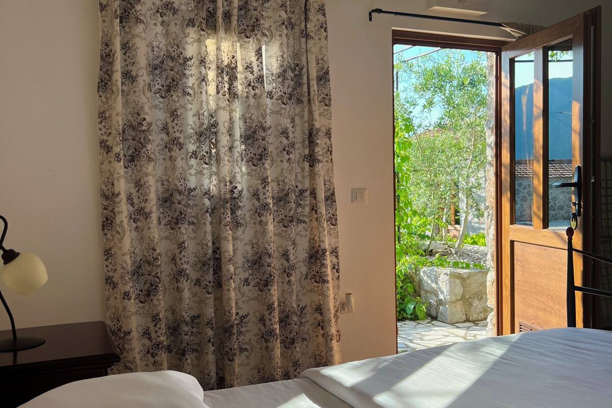 A bedroom with a bed, a nightstand, a lamp, and a view of the outdoors through an open door.