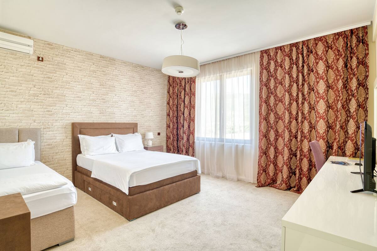 A hotel room with a brick wall, a large bed with a wooden headboard, a desk with a computer, and a window with patterned curtains.