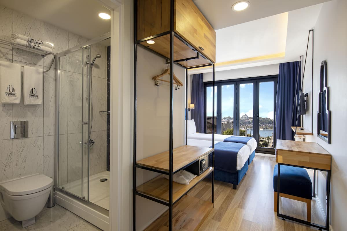 A modern hotel room with a glass shower enclosure, a wooden desk, a bed with blue bedding, and a view of the city.