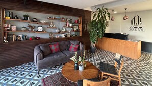 Een receptiegedeelte met een houten receptiebalie, een bank met kleurrijke kussens, een grote potplant en een wand met planken waarop diverse items staan.