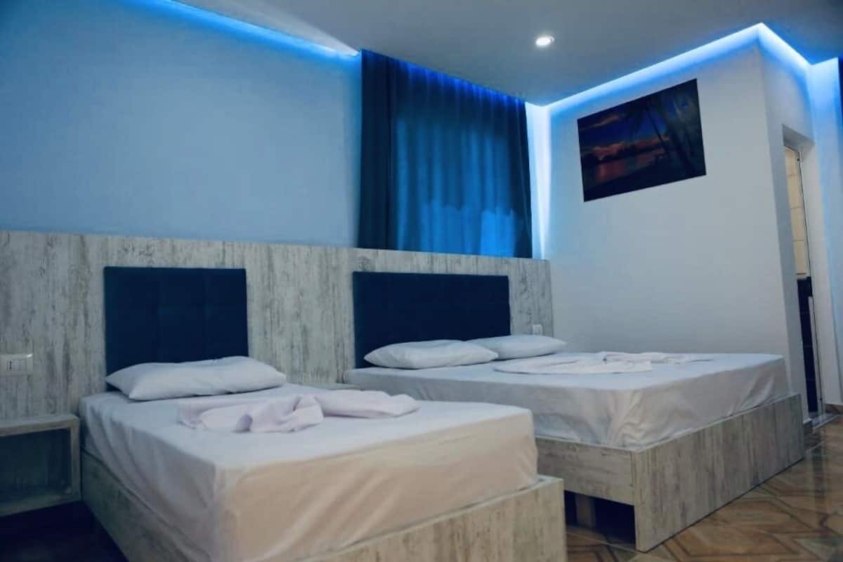 A hotel room with two beds, a large window with blue curtains, and a painting on the wall.