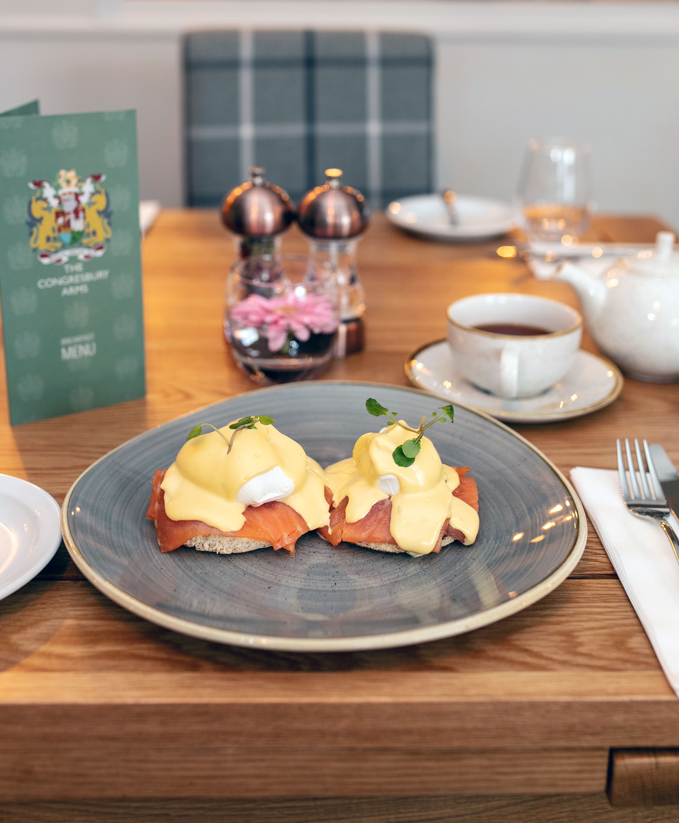 A table with a plate of eggs benedict, a menu, a teapot, and a glass of water.