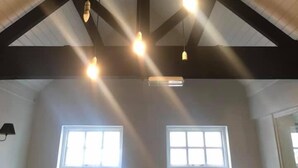 A room with a high ceiling, exposed beams, and pendant lighting. There are two windows providing natural light. - The Congresbury Arms (Bristol)