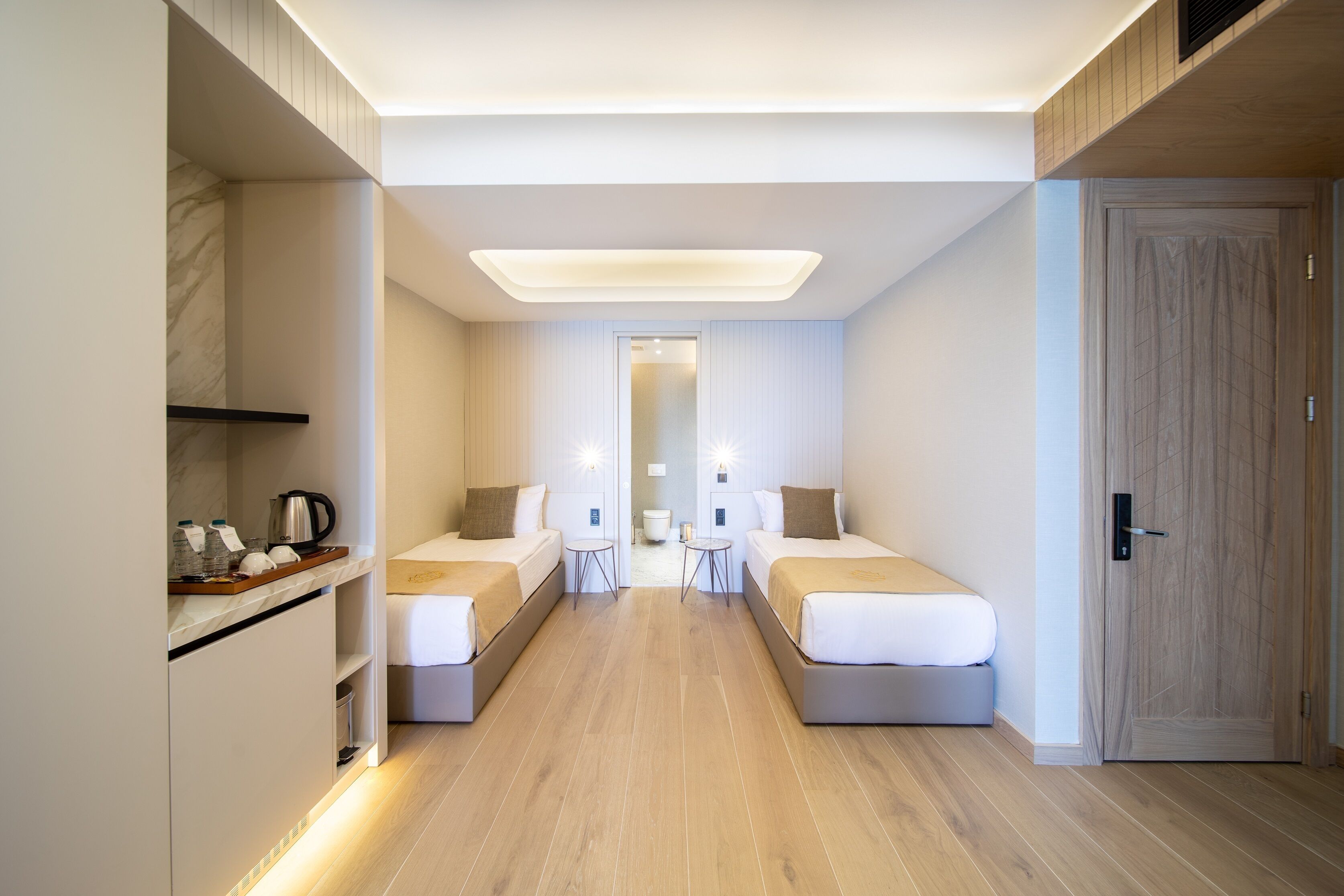 A modern hotel room with two beds, a small table, and a kitchenette area.