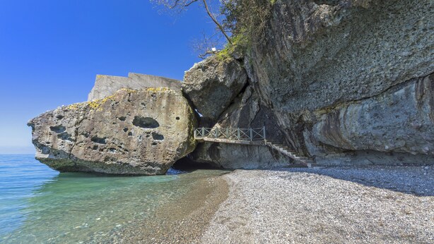 Une côte rocheuse bordée d’un ciel bleu azur, une plage de galets et un pont en bois menant à une falaise.