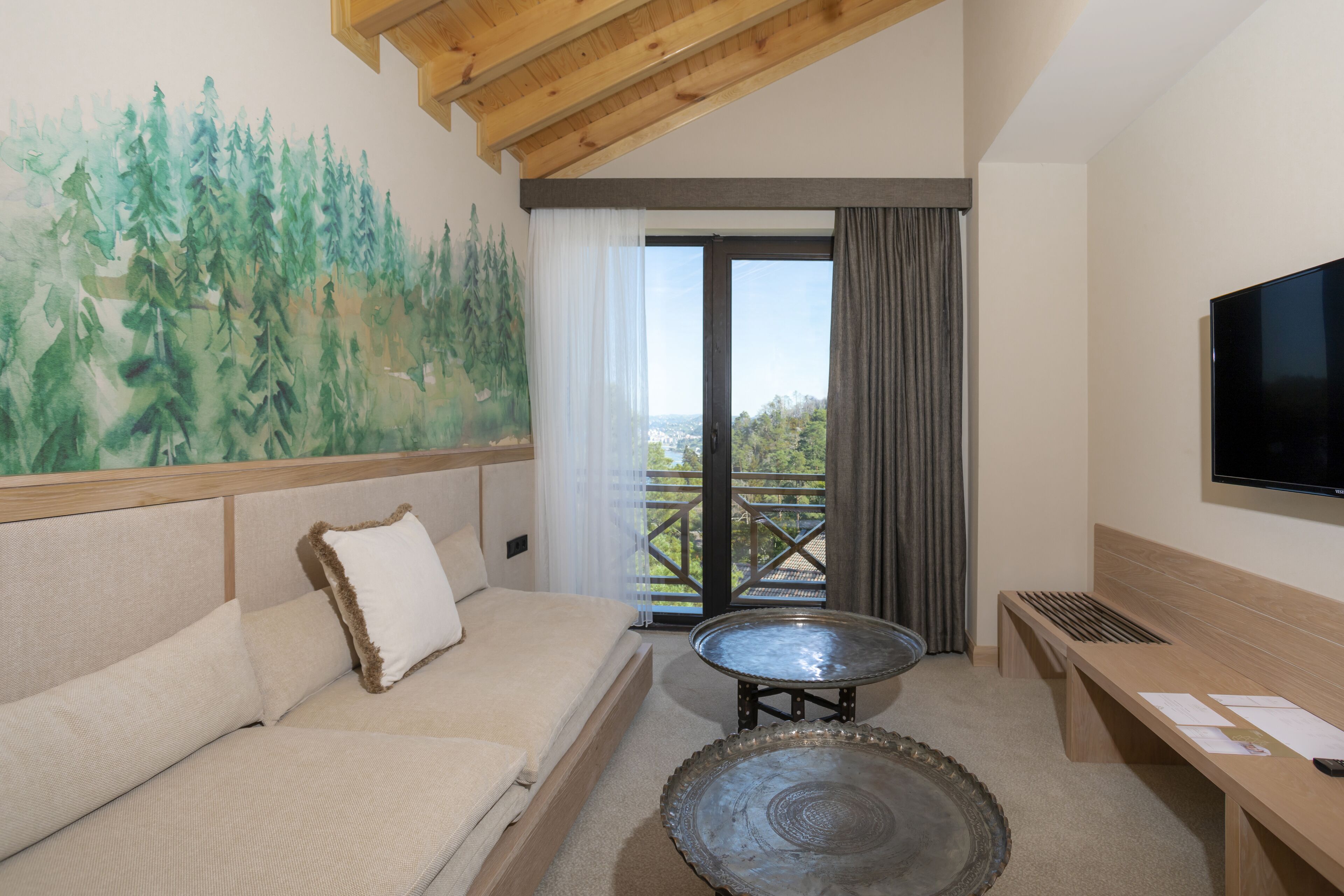 A room with a sofa, a small table, a bench, a TV, and a painting of trees.