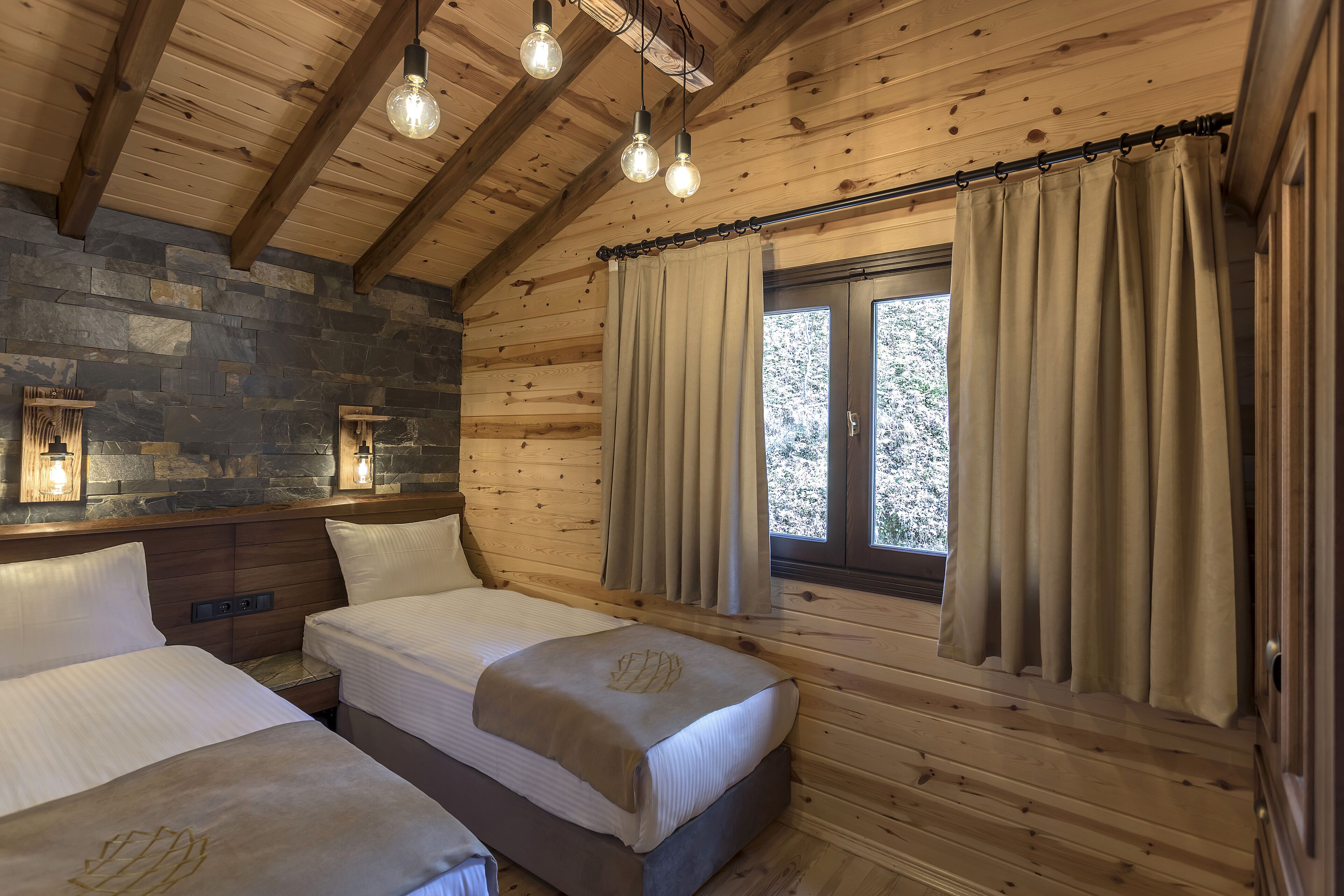 A wooden cabin bedroom with two beds, stone wall, and a window with curtains.