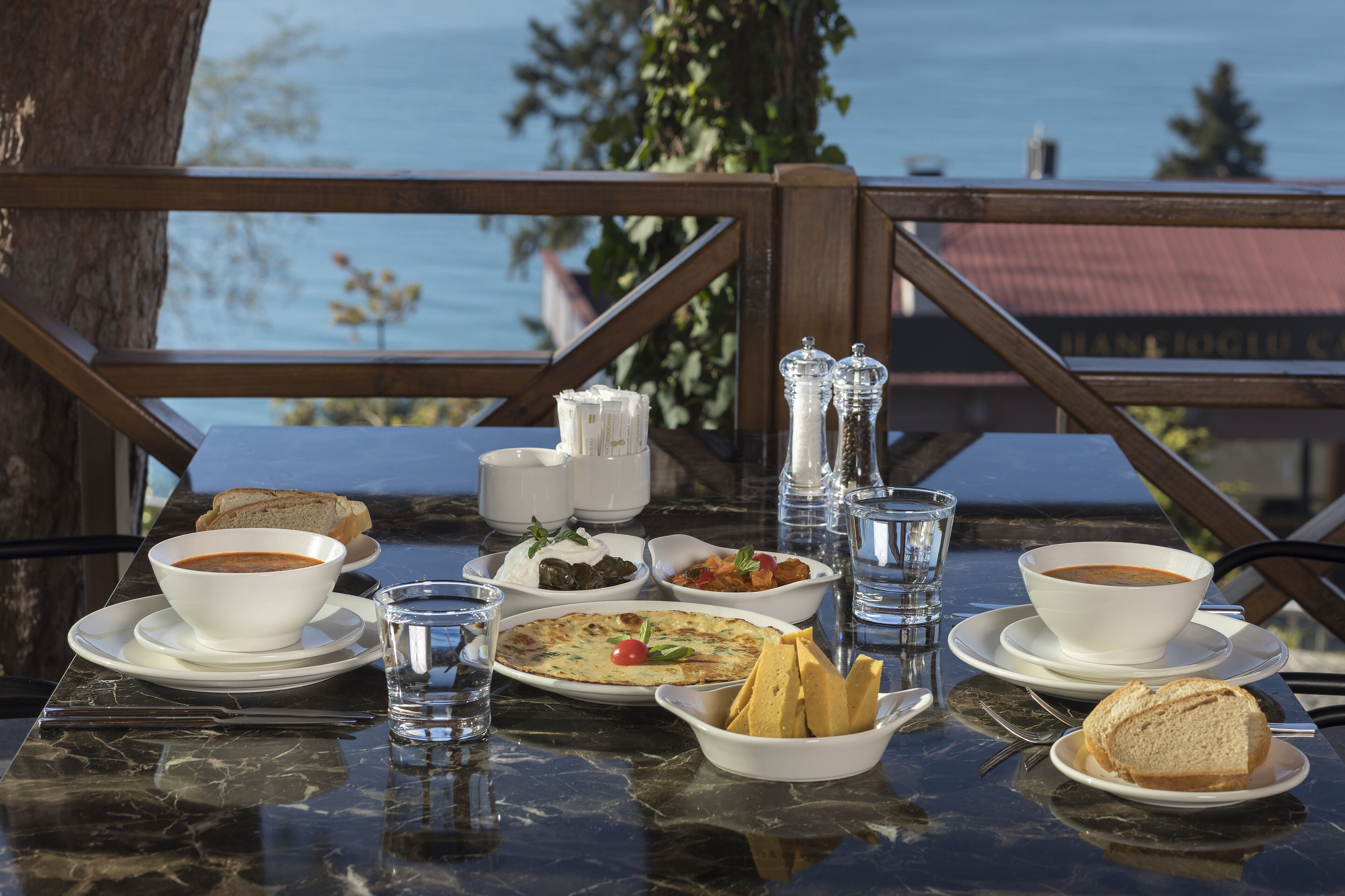 A table set with various dishes including soup, bread, and a salad, with a view of the sea in the background.