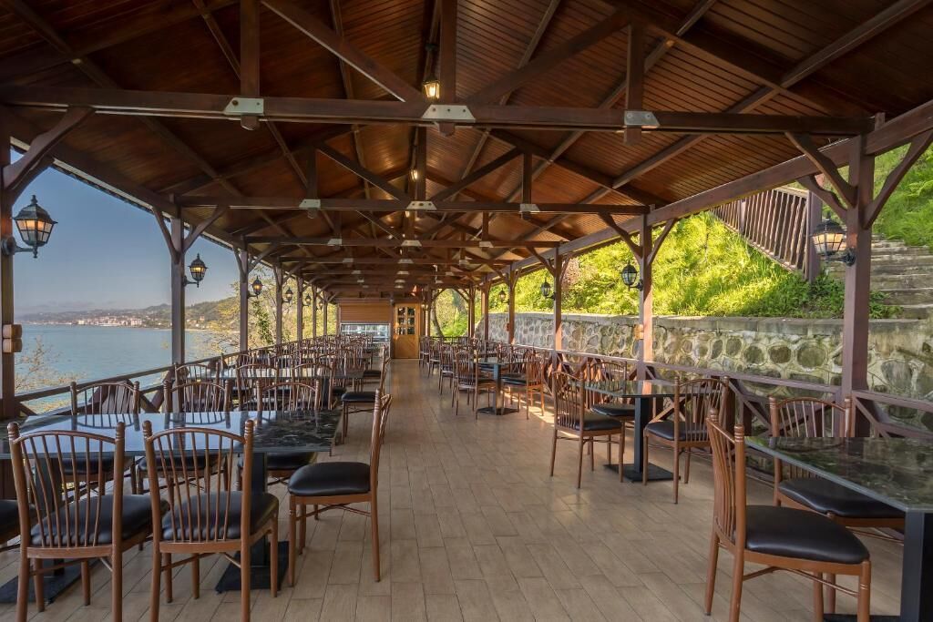 Un área de comedor al aire libre con mesas y sillas de madera, paredes de piedra y vistas al mar.