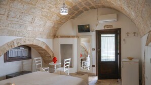 Una stanza con pareti in pietra, un letto, un tavolino, due sedie, un frigorifero e una televisione appesa al muro.