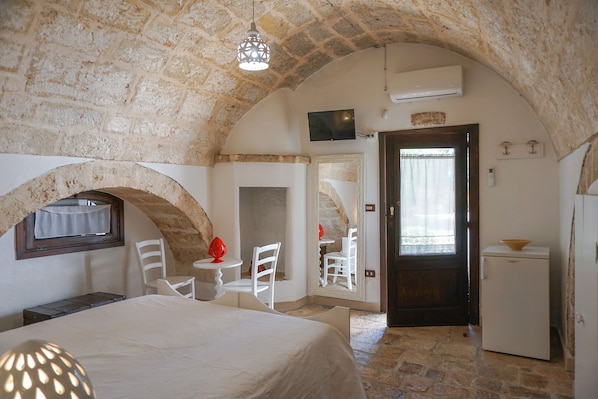 A stone-walled room with a bed, a small table, two chairs, a refrigerator, and a television on the wall. - Dal Canonico (Mola di Bari)
