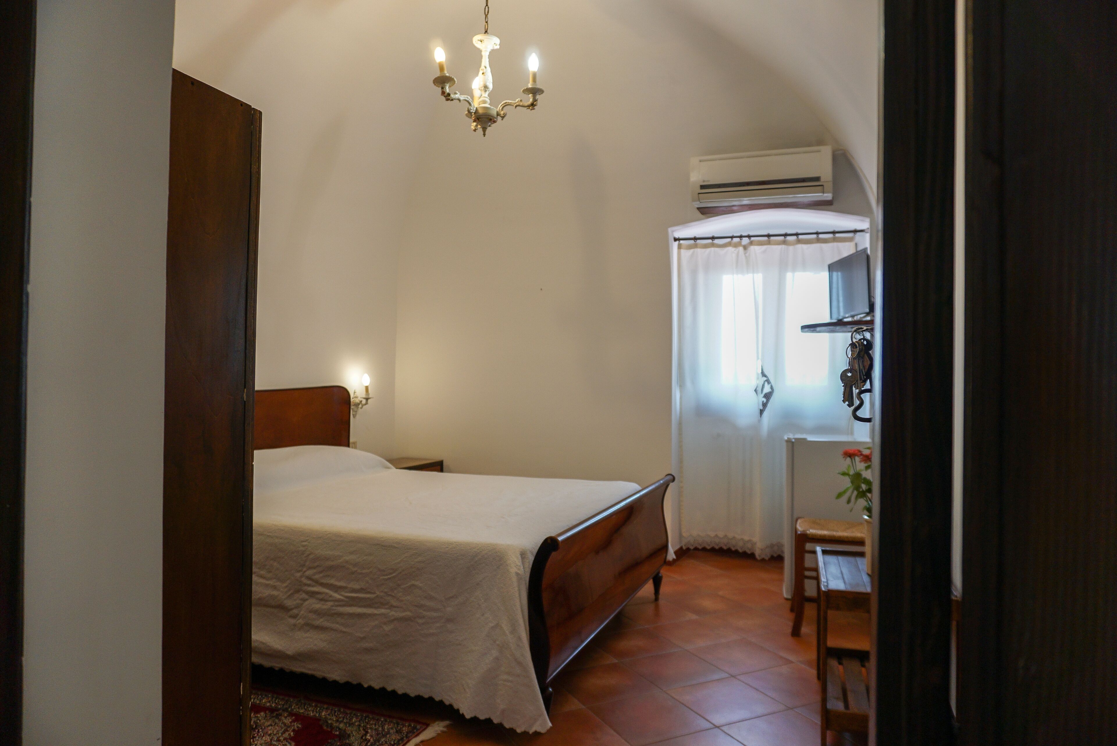 A bedroom with a wooden bed, a nightstand, a small table with a chair, a chandelier, and an air conditioning unit.